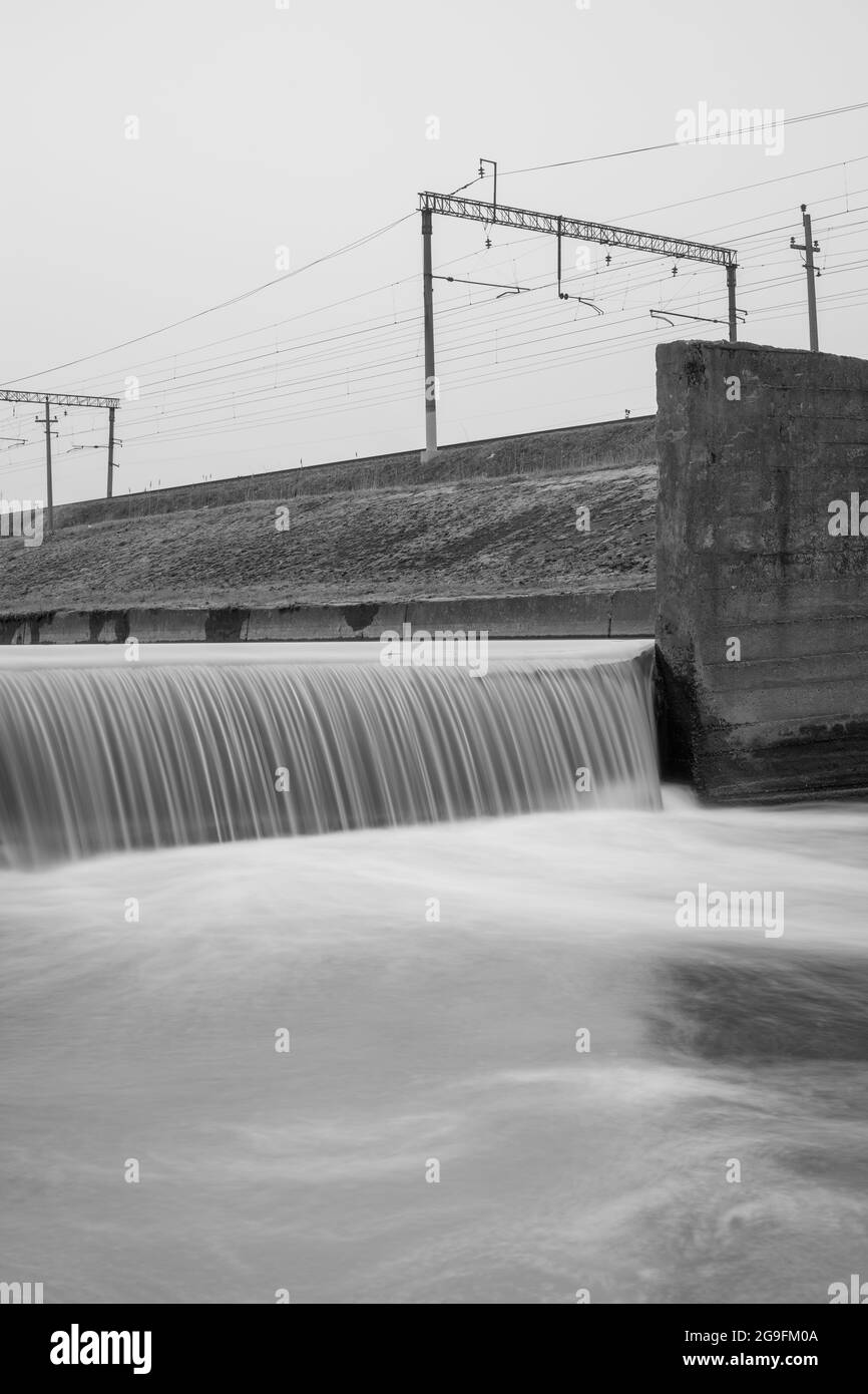 Dislivello in un canale di cemento vicino a binari ferroviari. Fotografia in bianco e nero a lunga esposizione. Foto Stock