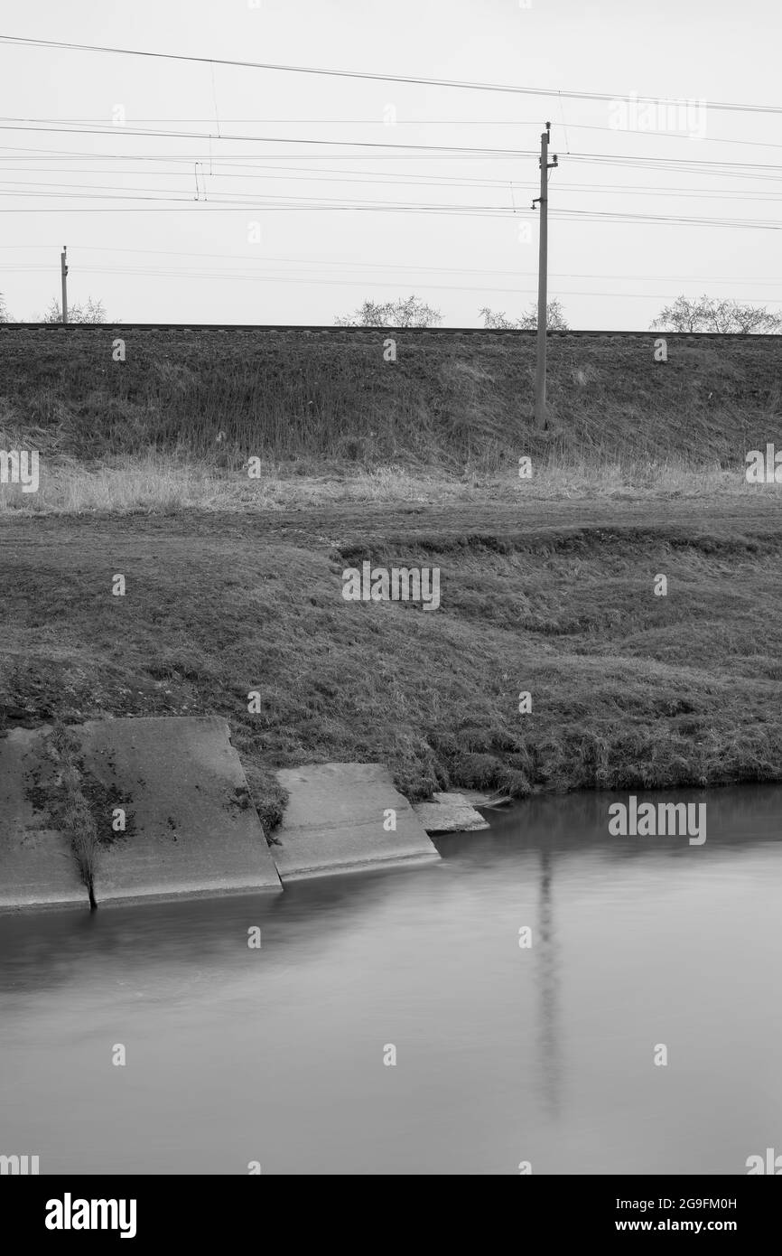 Canale di drenaggio dell'acqua vicino a binari ferroviari. Fotografia in bianco e nero a lunga esposizione. Foto Stock