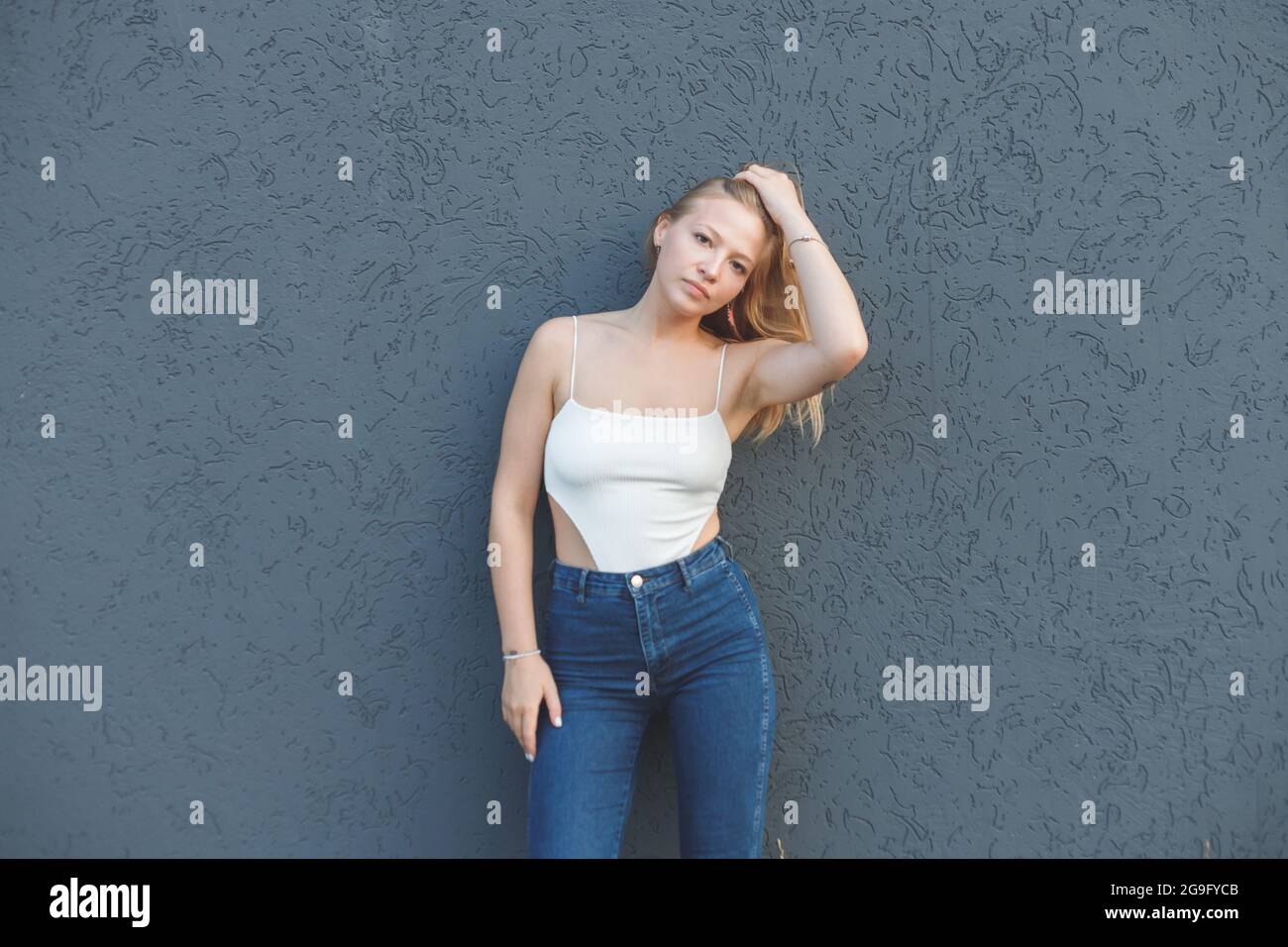 giovane donna bionda con la t-shirt bianca senza maniche e jeans blu che si erono su sfondo grigio della parete Foto Stock