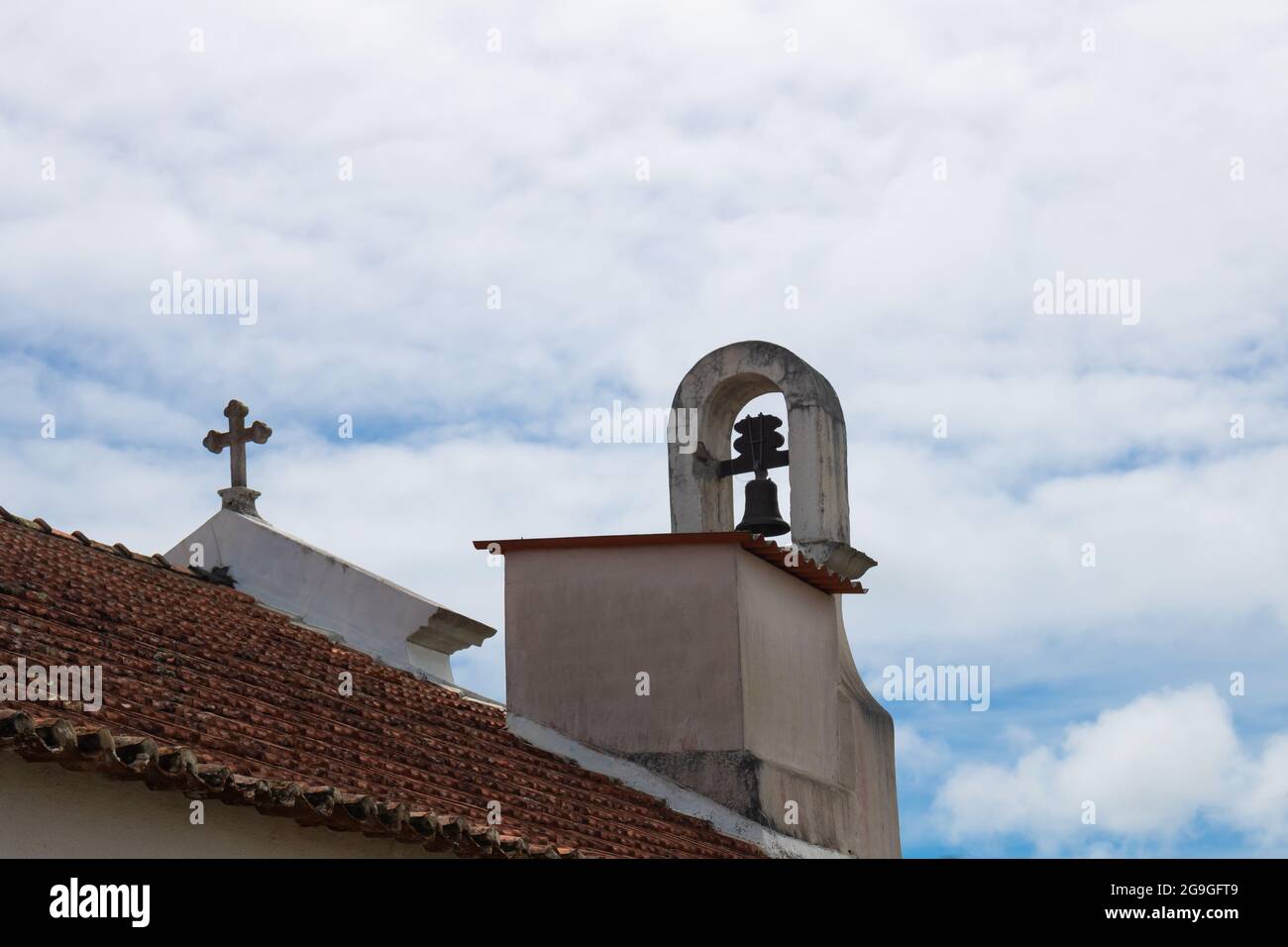 Particolare del tetto di una cappella con mattonelle arancioni, una croce cristiana e campanile contro il cielo blu nuvoloso in Portogallo Foto Stock