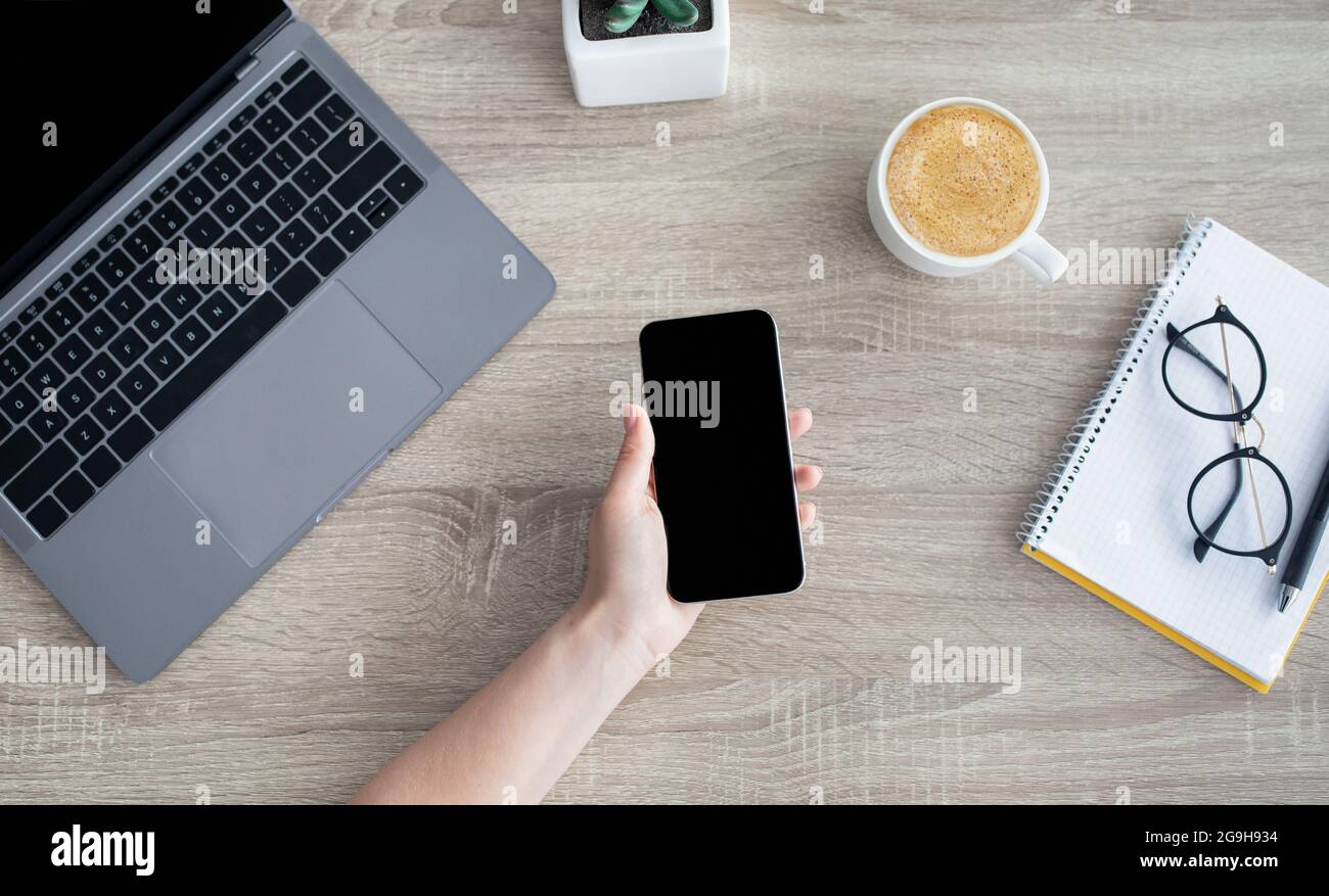Cellulare femminile con custodia a mano con schermo nero vuoto per il tuo messaggio di testo pubblicitario o contenuti promozionali, seduto sul posto di lavoro con computer portatile, in alto Foto Stock