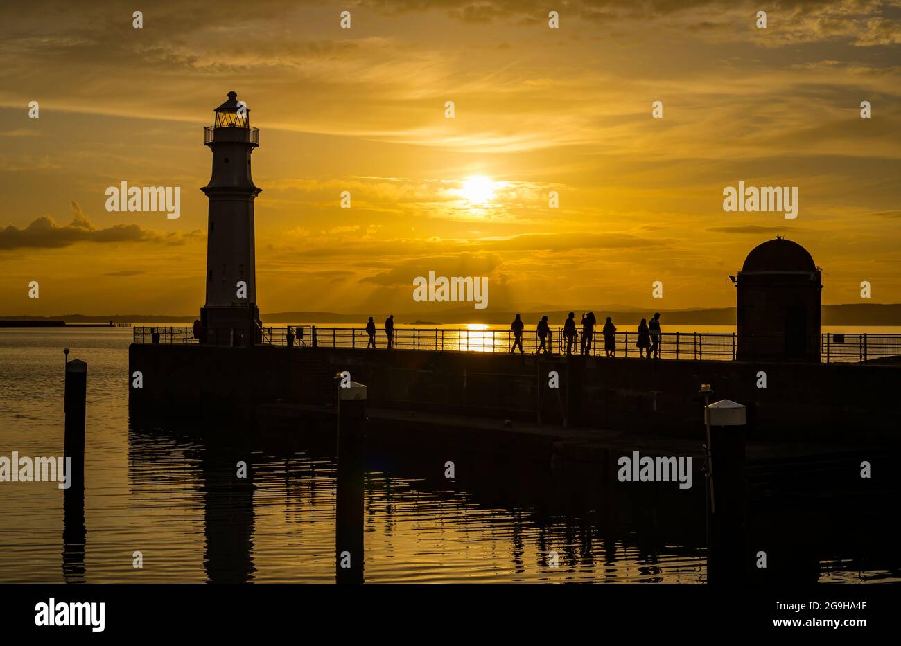 Le persone si sono sfrecciate sul molo del porto di Newhaven e sul faro in un colorato cielo arancione al tramonto, Edimburgo, Scozia, Regno Unito Foto Stock