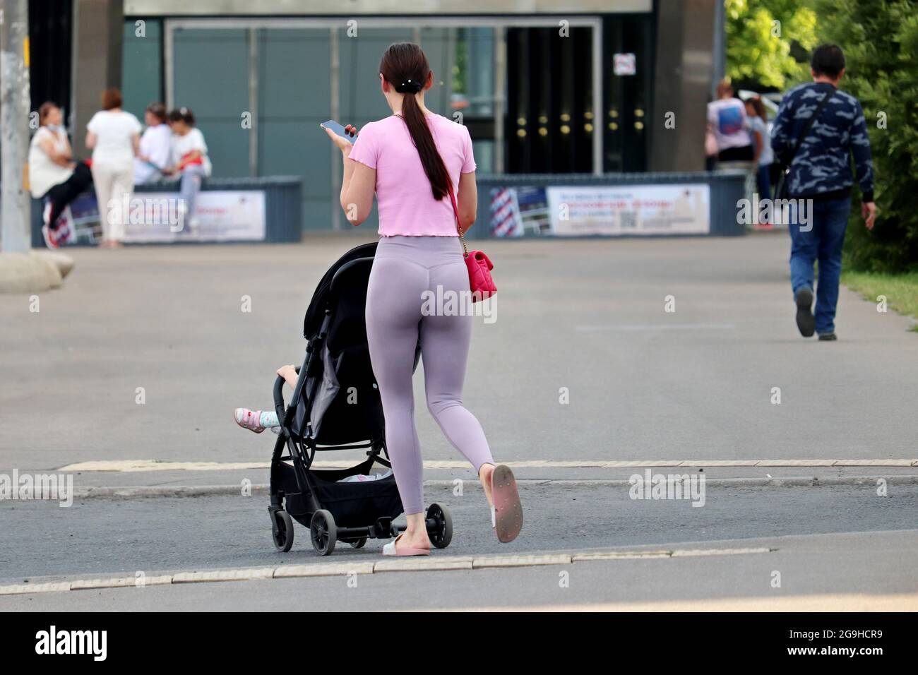Donna sottile in leggings con un passeggino che attraversa la strada della città con uno smartphone in mano. Concetto di maternità, mamma da passeggio con pRAM Foto Stock