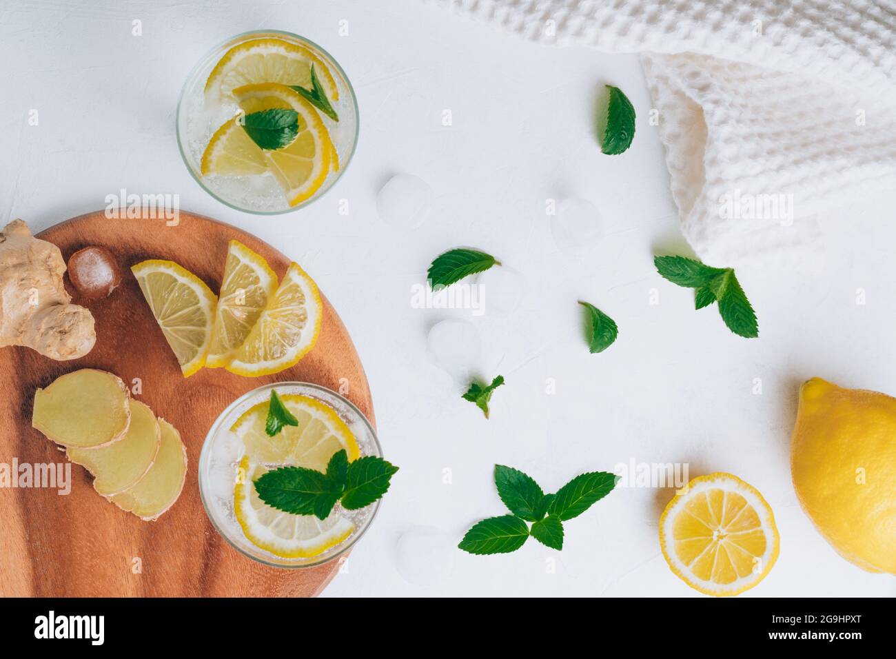 Due bicchieri con limonata e ingredienti per cucinare. Zenzero, limone, menta, ghiaccio su superficie bianca. Vassoio di legno piatto. Vista dall'alto Foto Stock