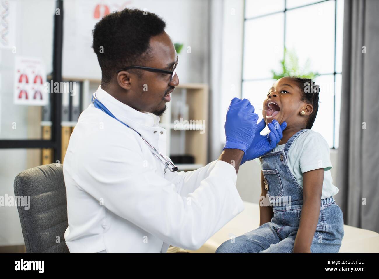 Bella bambina afro-americana seduta sul divano presso l'ufficio del medico, mentre bel medico maschio nero che esamina la gola e la bocca con la spatola. Concetto di Pediatria Foto Stock