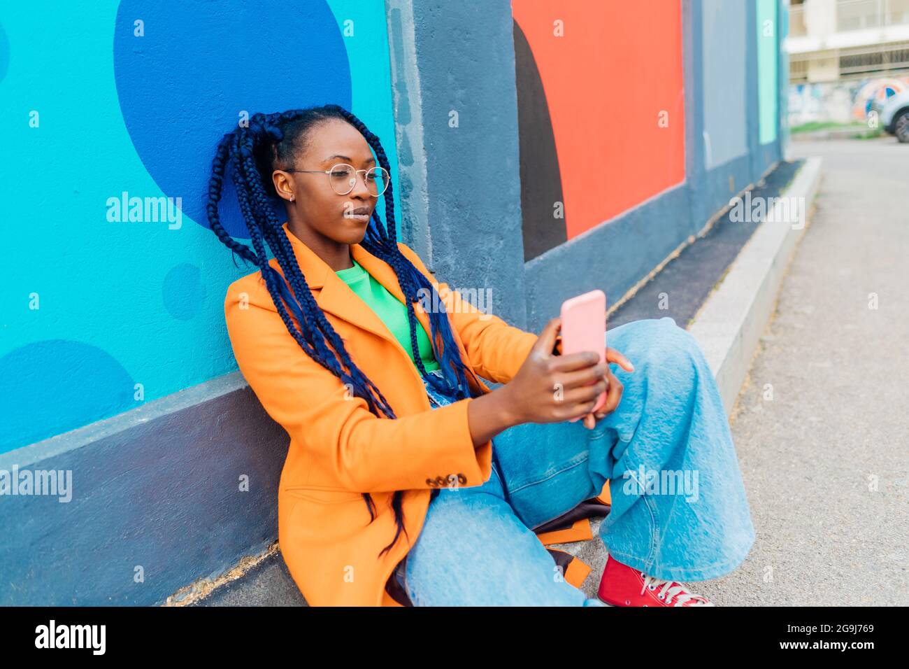 Italia, Milano, Donna con trecce seduta da parete colorata, utilizzando smartphone Foto Stock