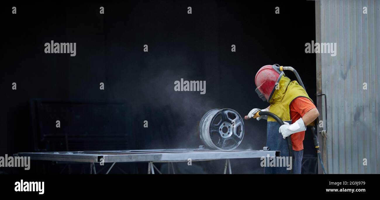 Uomo caucasico vestito con speciali dispositivi di protezione utilizzando sabbiatrice per la pulizia dei dettagli metallici sul lavoro. Concetto di persone e riparazione. Foto Stock