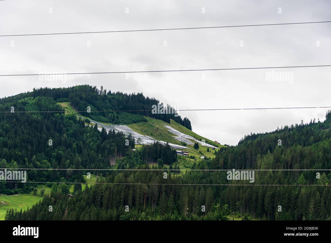 Parco solare, centrale fotovoltaica situata su un pendio di montagna nelle Alpi visto attraverso la linea elettrica aerea ad alta tensione. Foto Stock
