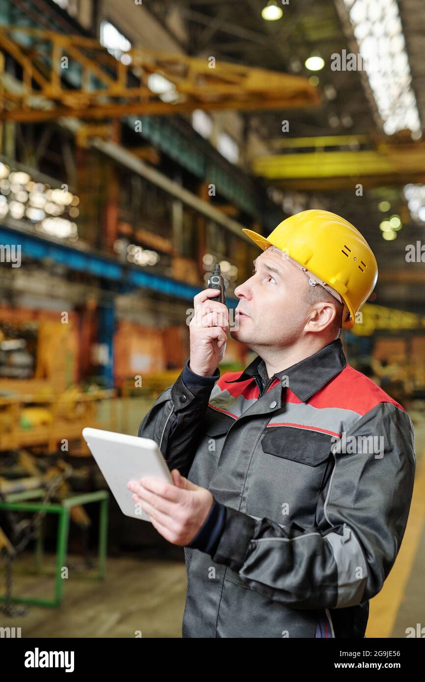 Ritratto di lavoratore manuale maturo in casco e abbigliamento da lavoro con la fotocamera mentre si utilizza un tablet pc in fabbrica Foto Stock