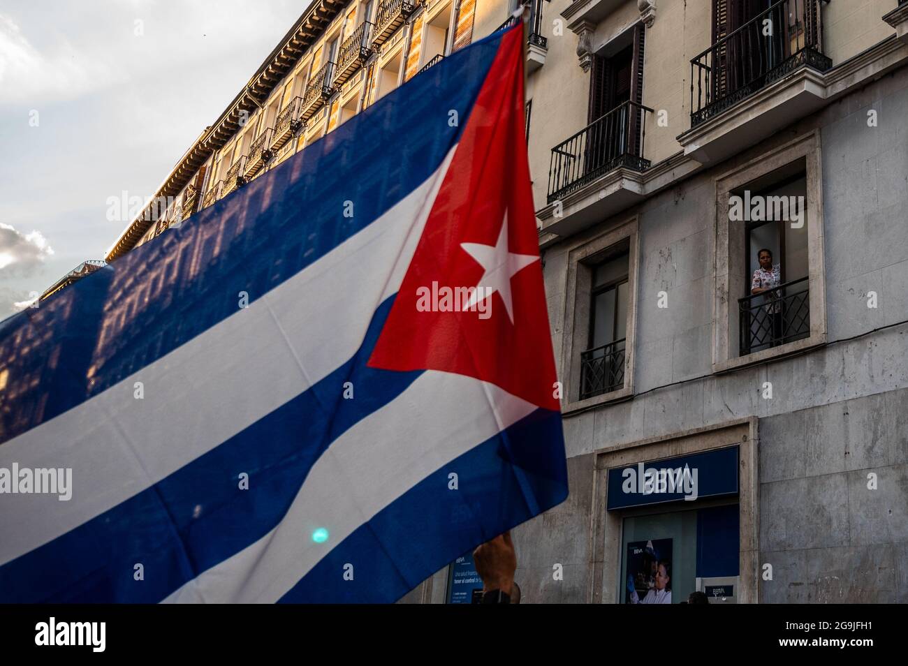 Madrid, Spagna. 26 luglio 2021. Una donna guarda da un balcone come manifestanti con bandiere passano durante una manifestazione contro l'embargo economico statunitense su Cuba, in coincidenza con il 26 luglio, il giorno in cui la rivoluzione cubana è iniziata nel 1953, 68 anni fa. Credit: Marcos del Mazo/Alamy Live News Foto Stock