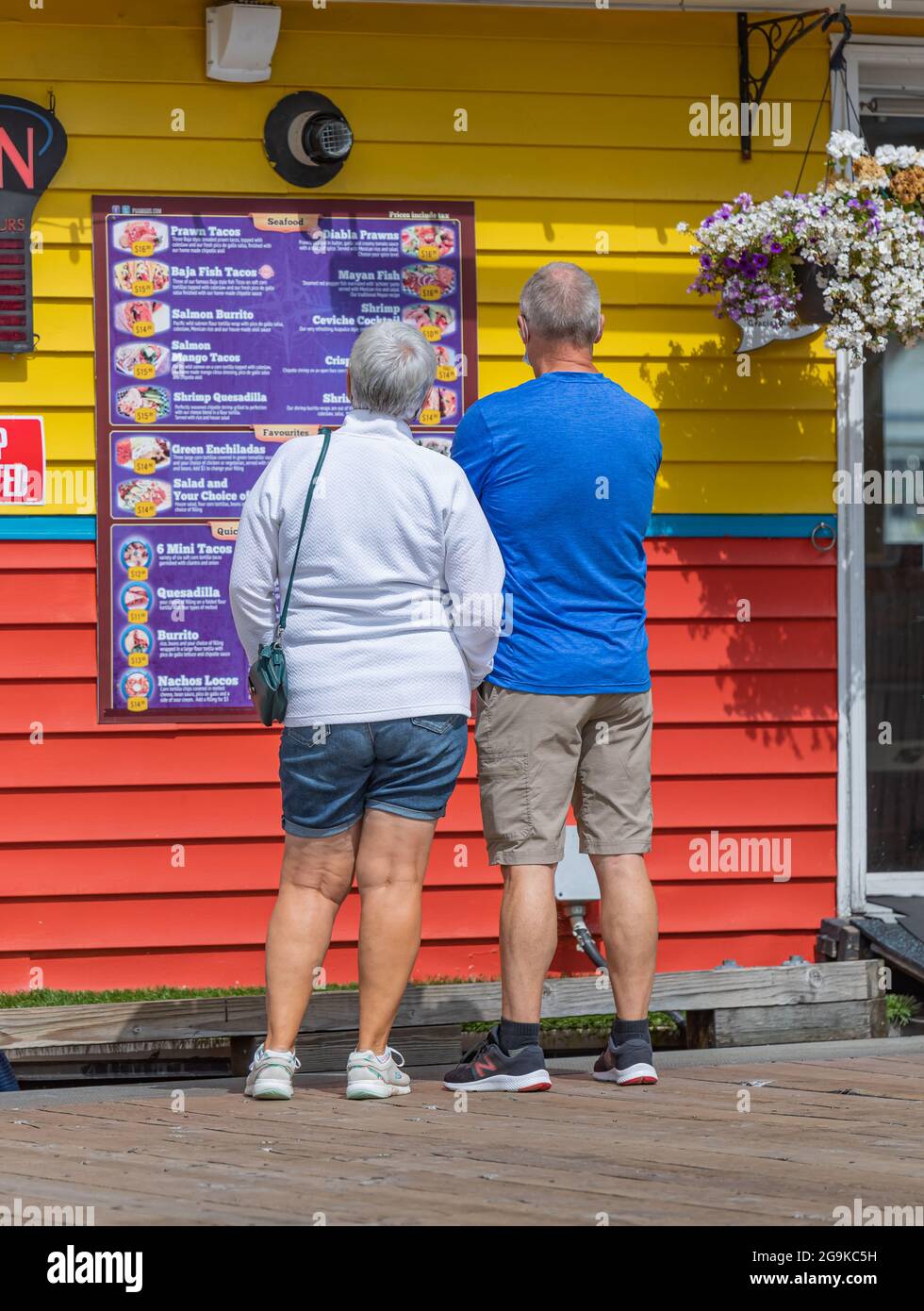La coppia anziana si trova sulla strada e guarda al menu della caffetteria all'esterno. Fisherman Wharf, Victoria, BC, Canada-Luglio 23,2021. Foto di strada, t Foto Stock