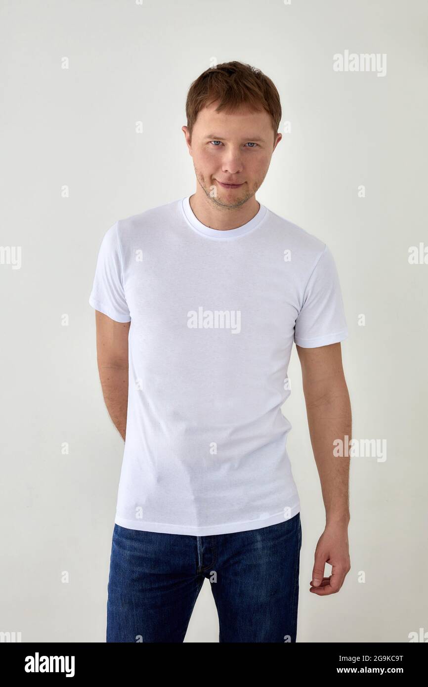 Giovane uomo positivo in jeans con la mano dietro e la t-shirt bianca in piedi su sfondo bianco e guardando la macchina fotografica Foto Stock