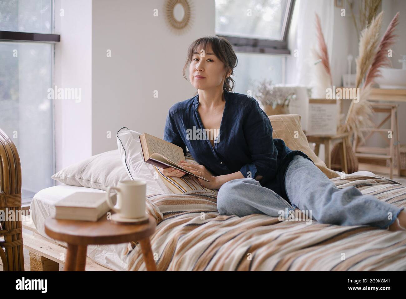 Bella giovane donna asiatica che legge un libro. Godendo di un momento tranquillo e rilassante ambiente in casa accogliente Foto Stock