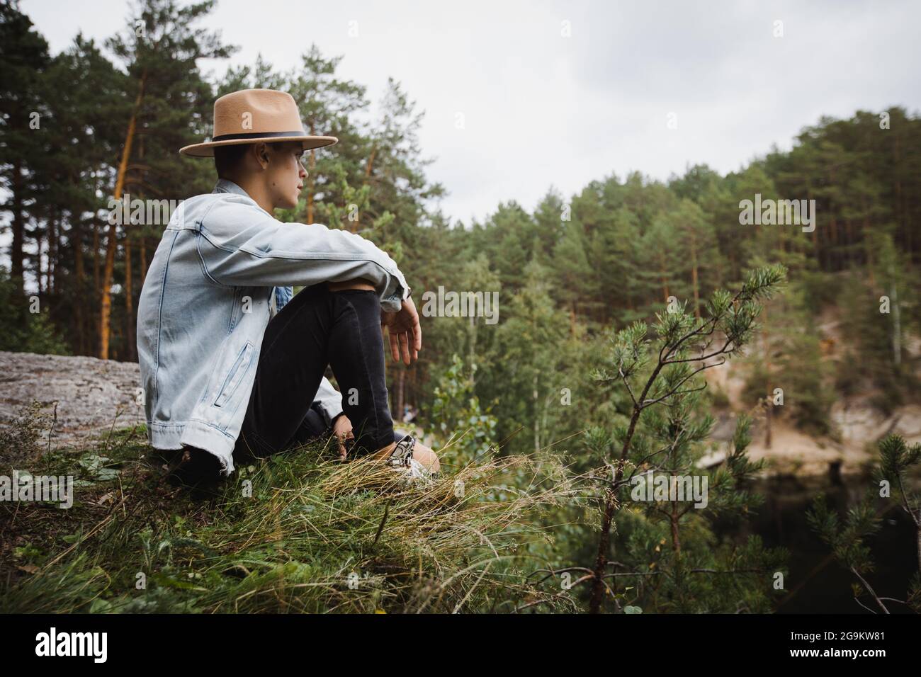 Vista laterale basso angolo di giovane viaggiatore maschile seduto sul bordo di pietra e ammirando il paesaggio del lago circondato da foreste di conifere Foto Stock
