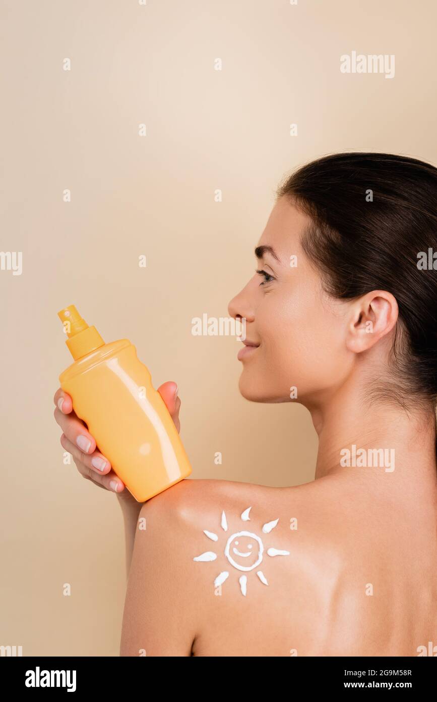donna sorridente con immagine di sole sorridente sulla spalla che tiene la protezione solare isolato su beige Foto Stock
