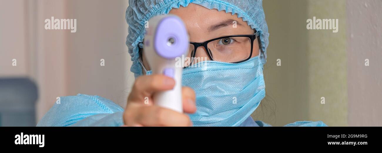 il banner con infermiere o medico in tuta medica monouso misura la temperatura corporea del paziente con un termometro medico a infrarossi. Pistola termometro Isom Foto Stock