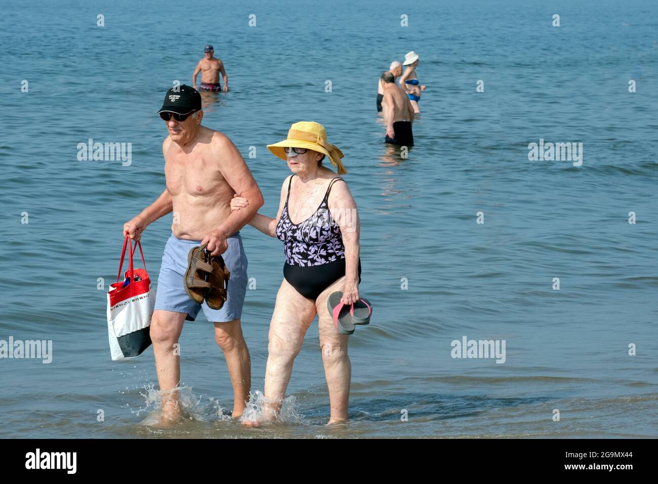 Una coppia più anziana, presumibilmente marito e moglie, cammina con il braccio lungo il litorale di Brighton Beach a Brooklyn, New York. Stanno tenendo i loro sandali Foto Stock