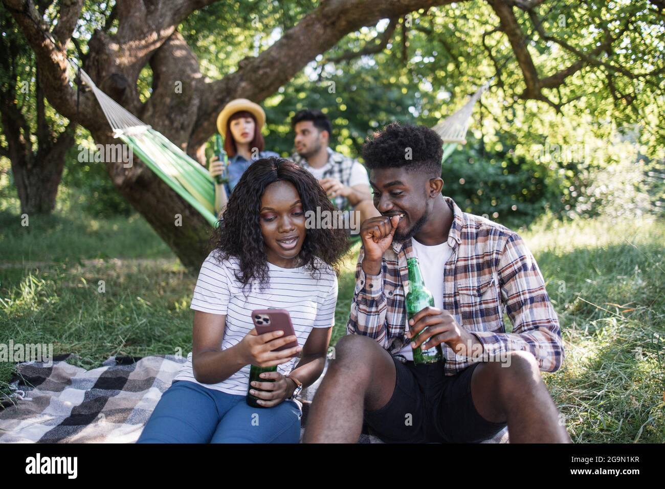 Coppia afroamericana che usa uno smartphone moderno mentre si siede insieme sulla plaid nel giardino estivo. Amici multirazziali che chiacchierano e bevono birra in amaca sullo sfondo. Foto Stock