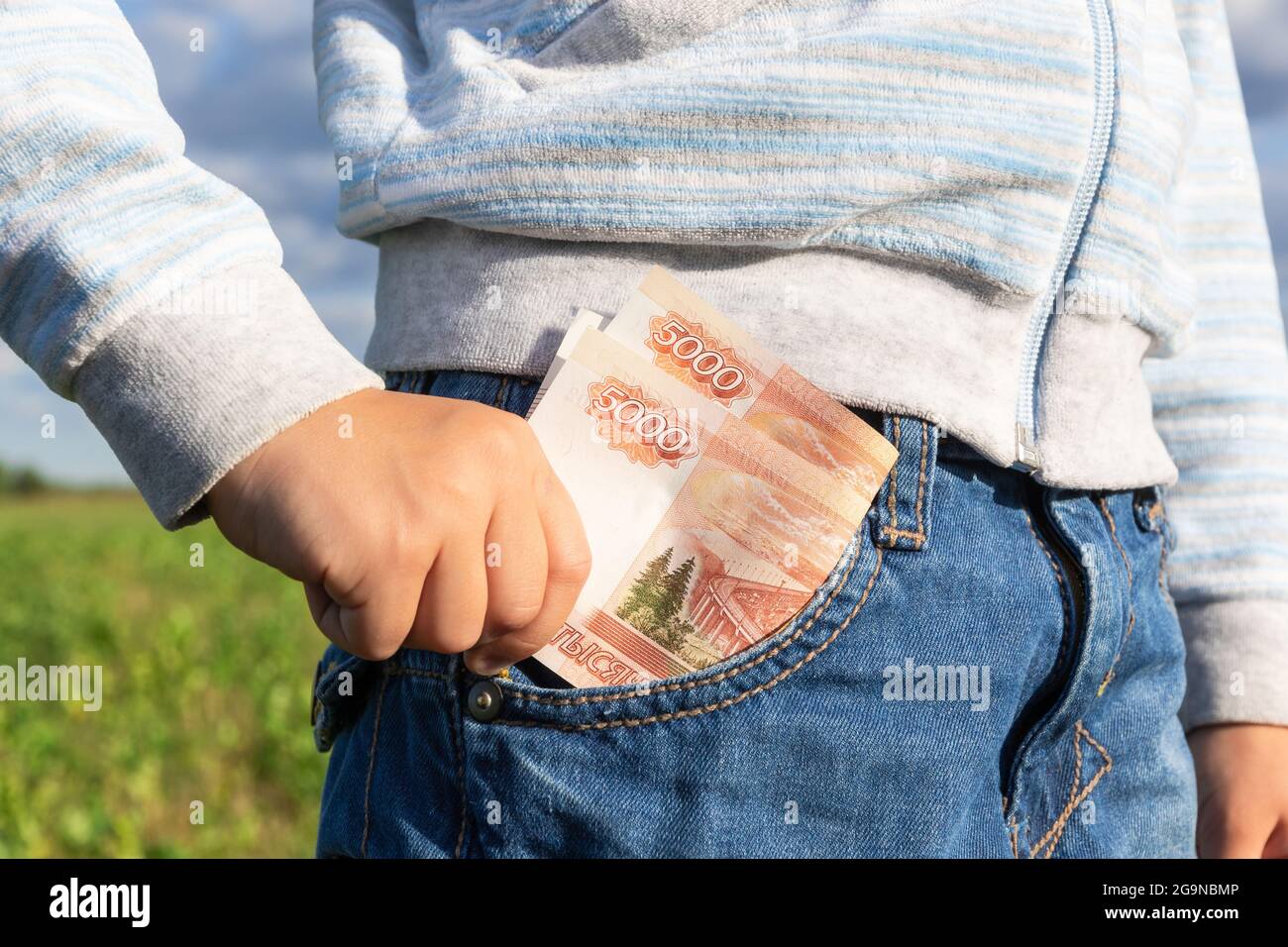 Una mano del bambino toglie i rubli di soldi di carta da una tasca dei  jeans su uno sfondo di cielo blu in una giornata estiva luminosa e  soleggiata. Messa a fuoco