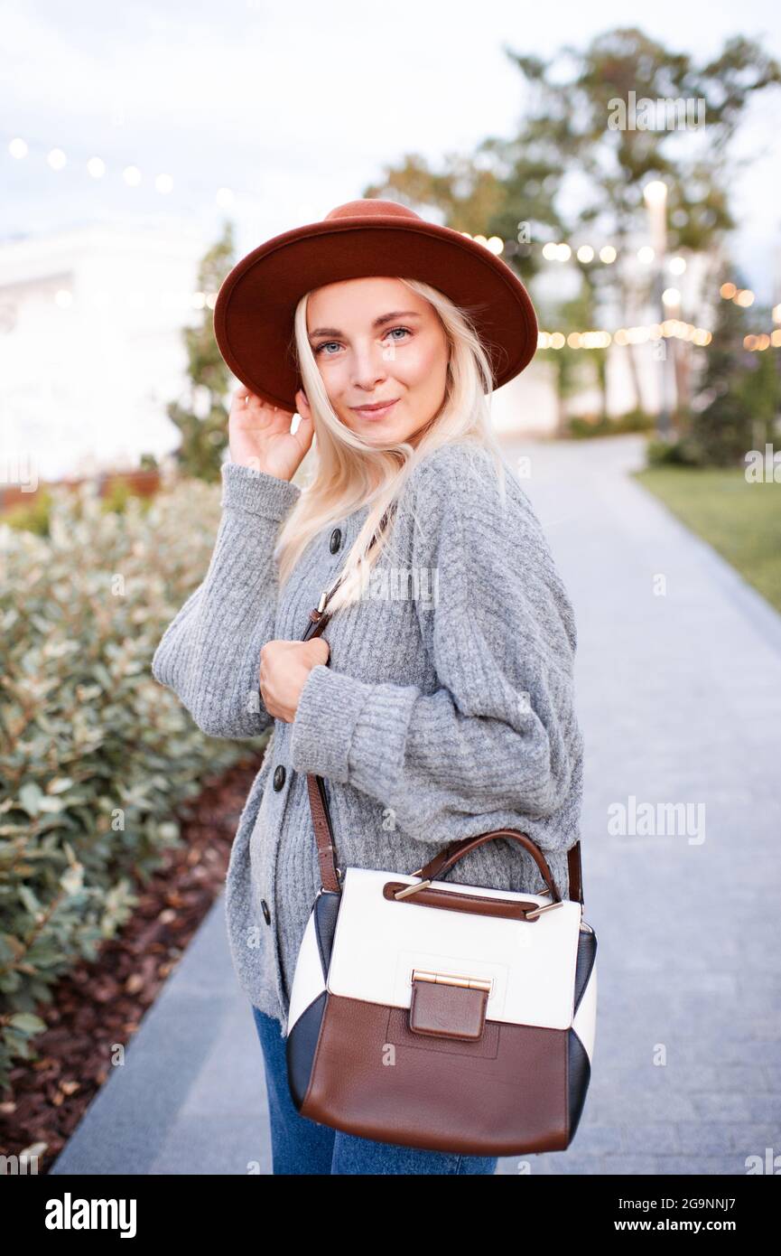 Felice elegante ragazza bionda 19-20 anni indossare cappello, maglia maglia maglia in lana jumper e tenere borsa in pelle camminare nel parco all'aperto. Guardando la fotocamera. Felicità. AUT Foto Stock