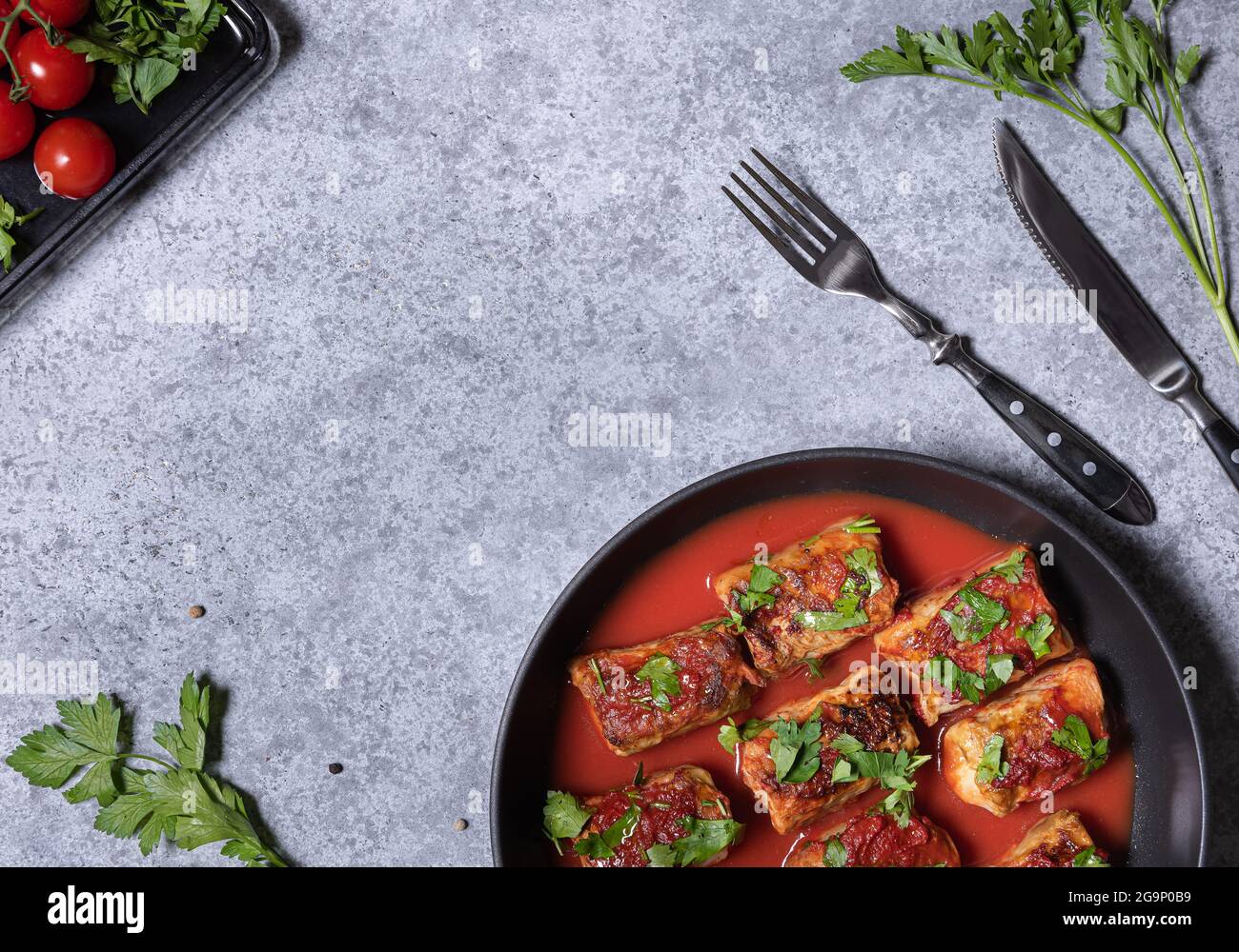 Involtini di cavolo, salsa di pomodoro e prezzemolo tritato in padella. Piatto con rotoli di cavolo, pomodori, prezzemolo, forchetta e coltello su texture grigie Foto Stock