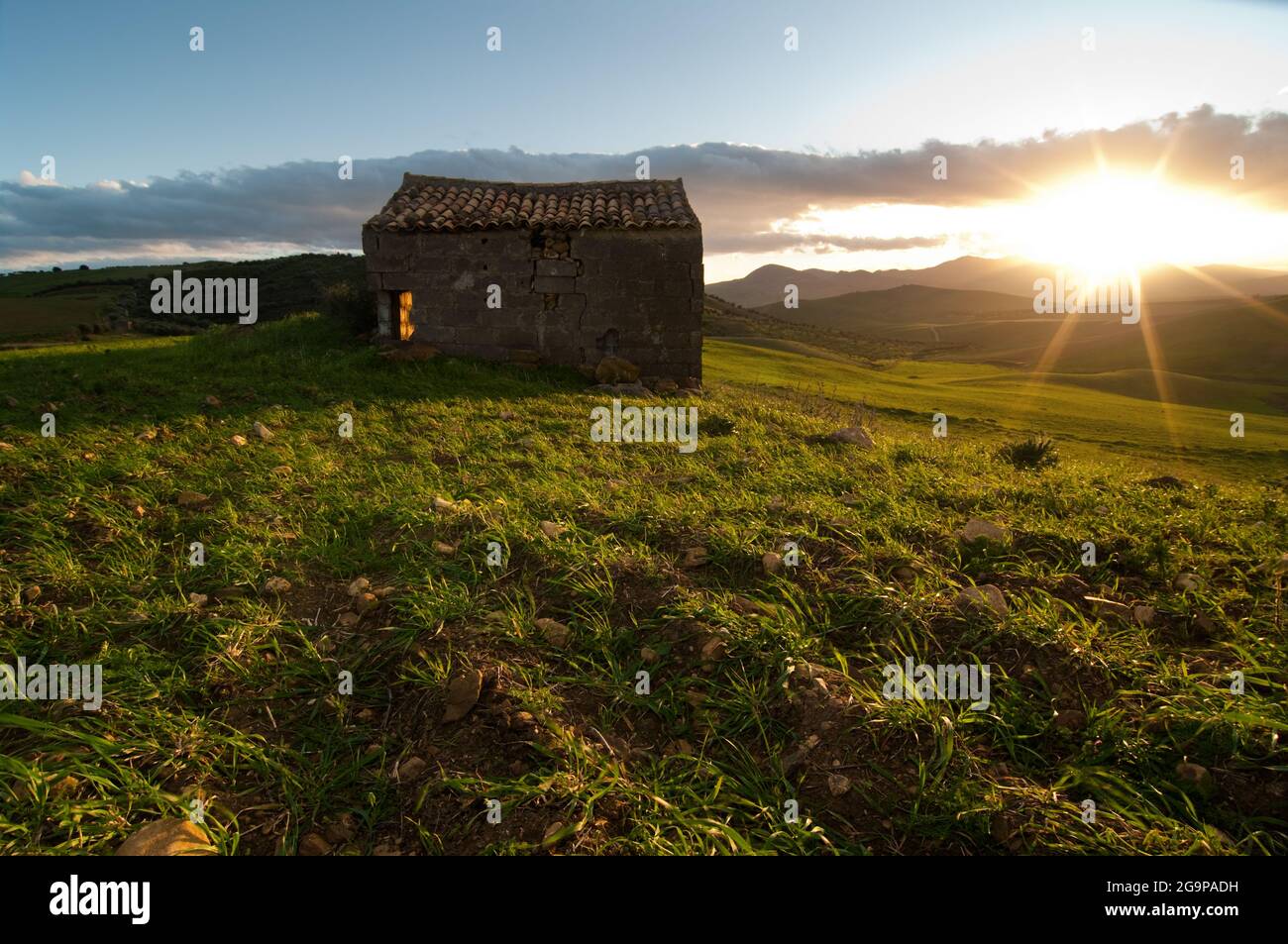 paesaggio di casolare abbandonato in campo di erba verde al tramonto con raggi luminosi Foto Stock