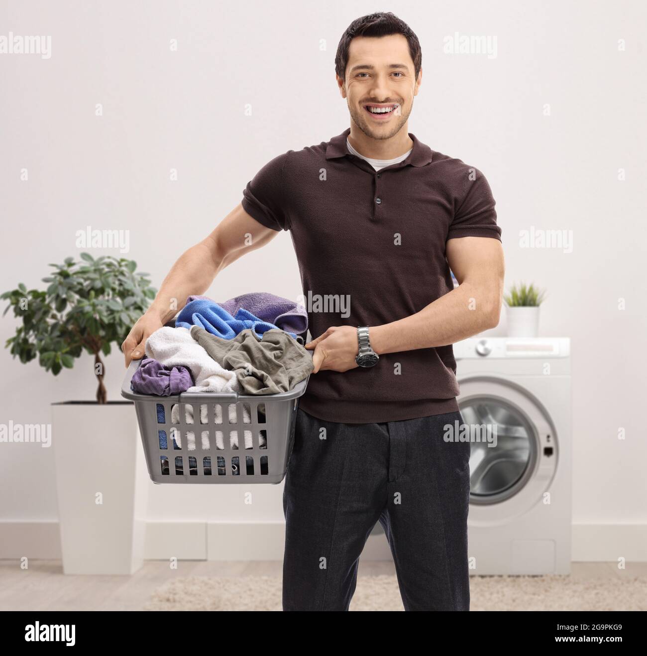 Un giovane sorridente con un cesto di biancheria pieno di vestiti in piedi in un bagno Foto Stock