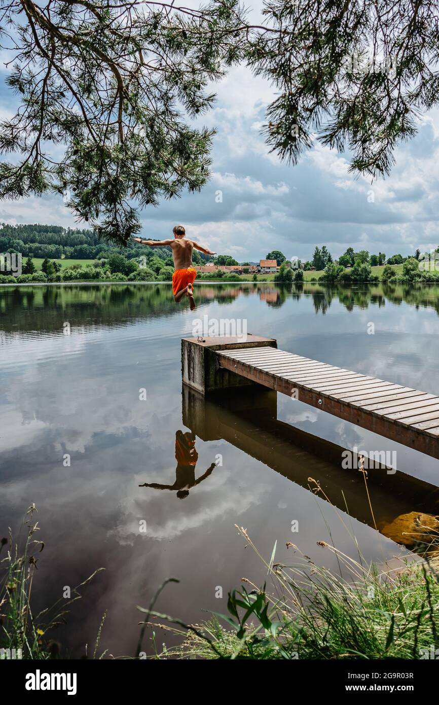 Vista posteriore dell'uomo che salta dal molo di legno con gioia in acqua, la natura riflessa in Lake.having estate divertimento da stagno.Travel vacanza, vacanza dei giovani Foto Stock