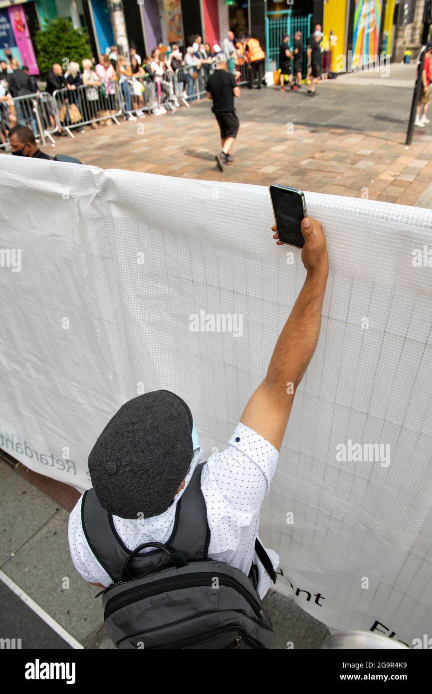 Glasgow, Scozia, Regno Unito. 27 luglio 2021. NELLA FOTO: Un uomo scatta una foto sul suo telefono con fotocamera che allunga il braccio sopra le scene per fermare la gente che cerca di vedere nel set di film. Scene del nuovo film The Flash che mostra batman stunt doppio sul set del film nel centro di Glasgow City Centre. Il nuovo campione d'incassi di Hollywood ha scelto il Glasgow City Centre per i suoi edifici dall'aspetto americano. Credit: Colin Fisher/Alamy Live News Foto Stock