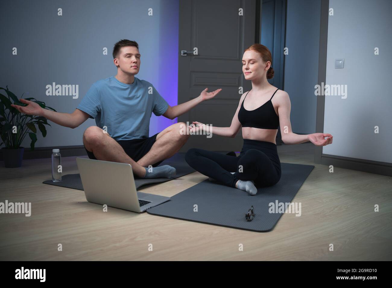 Un marito e una moglie trascorrono una lezione di yoga a casa seduti sul pavimento, meditano insieme davanti a un computer portatile. Foto Stock