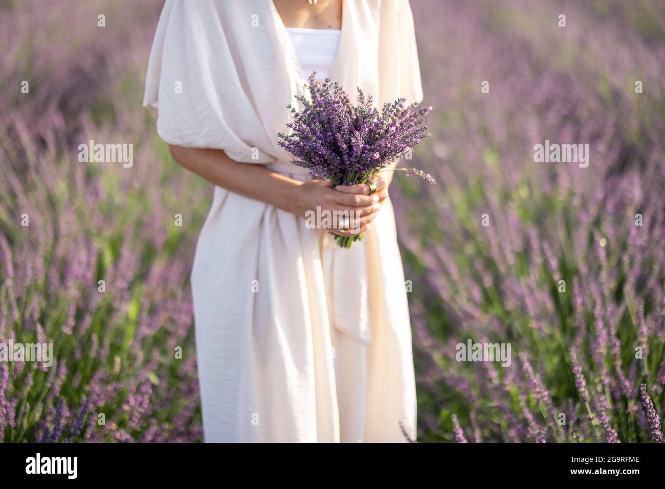 Donna in abito bianco e bouquet viola di fiori freschi di lavanda in mano su campo fiorito. Calma e concetto di consapevolezza. Godere di bellezza e profumo di natura in estate. Foto Stock