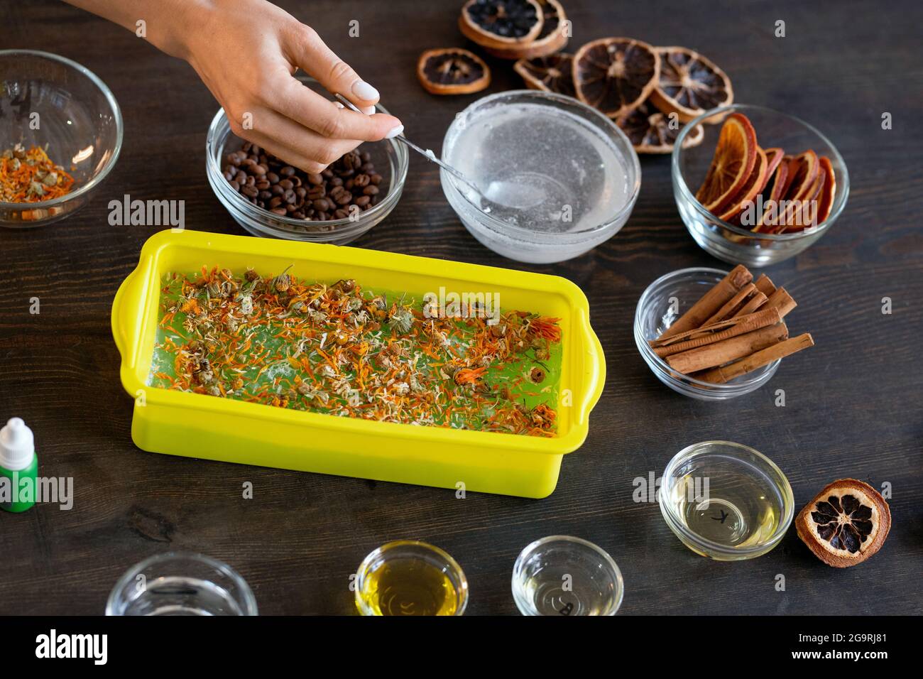 Mano di femmina che mette i fiori secchi di calendula in contenitore di silicone con massa di sapone fuso mentre mostra il suo amico come faccia i cosmetici naturali Foto Stock