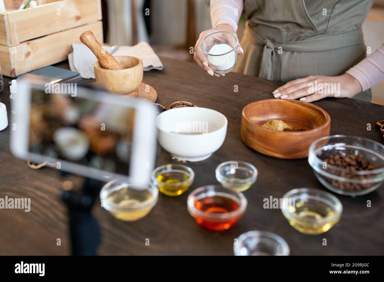 Smartphone sul supporto della fotocamera in piedi davanti al tavolo dove giovane donna che prepara ingredienti per il sapone naturale fatto a mano Foto Stock