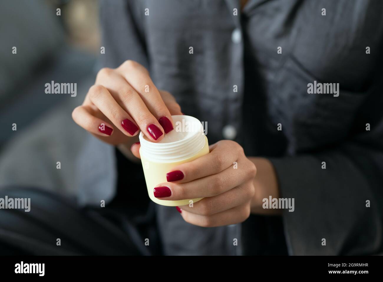 Giovane donna in pigiama grigio che applica un prodotto cosmetico naturale fatto a mano sulle sue mani mentre si siede a letto al mattino Foto Stock