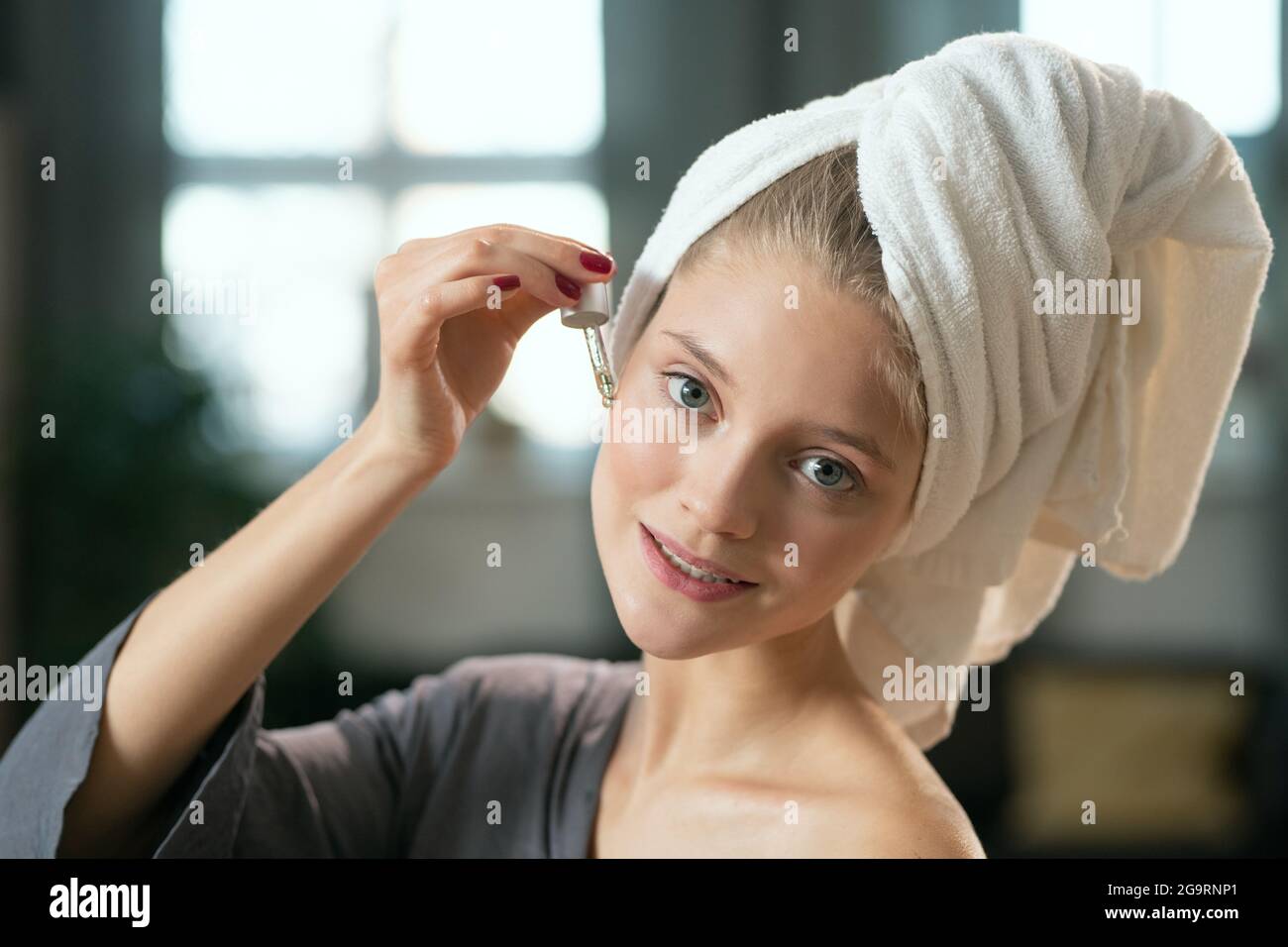 Giovane donna gorgeous felice in pigiama grigio seta che applica il cosmetic prodotto sul collo e sul corpo e godendo dopo bagno Foto Stock