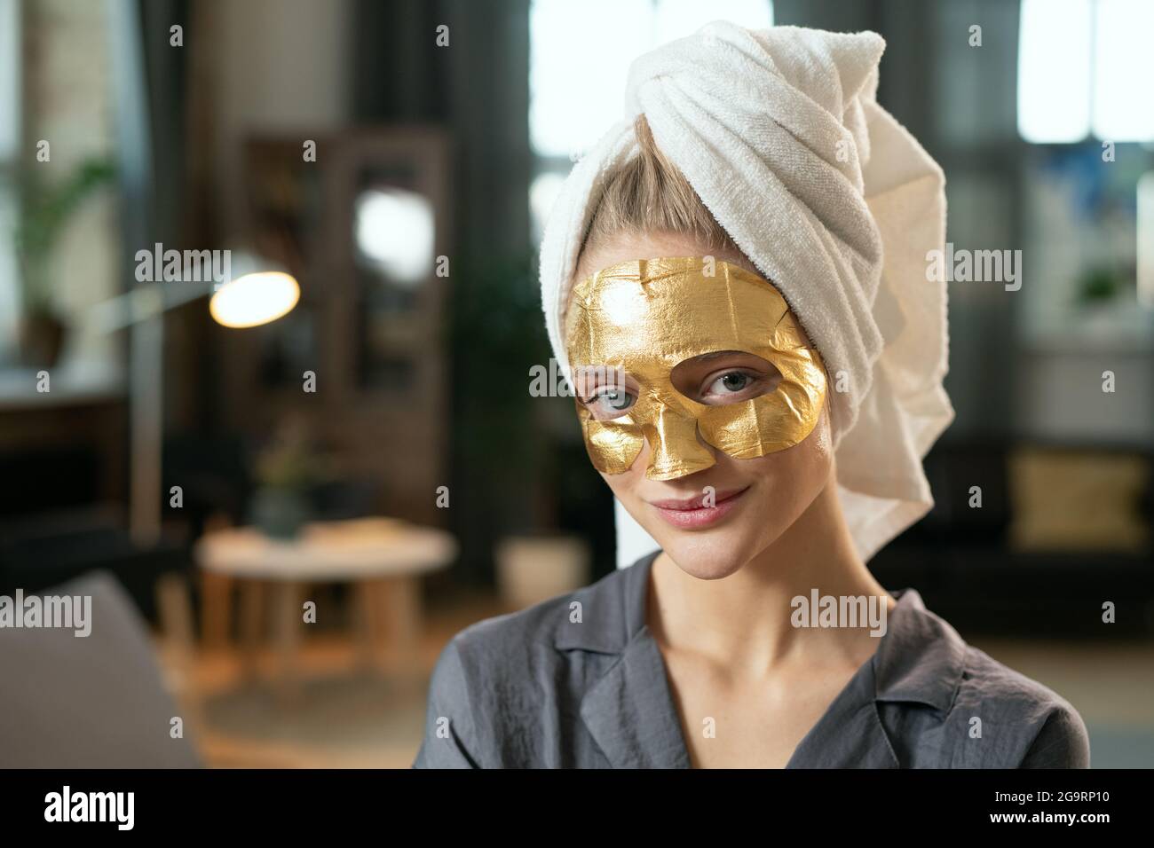 Giovane donna sorridente in pigiama grigio seta, asciugamano sulla testa e maschera cosmetica dorata sul viso rilassandosi a letto a casa e guardandovi Foto Stock
