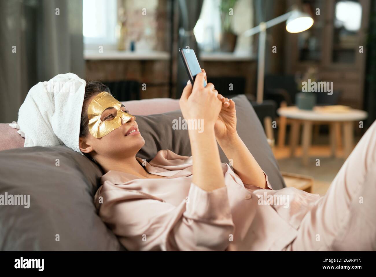 Giovane bella donna in pigiama di seta, asciugamano sulla testa e maschera cosmetica dorata sul viso rilassandosi a letto e facendo selfie Foto Stock