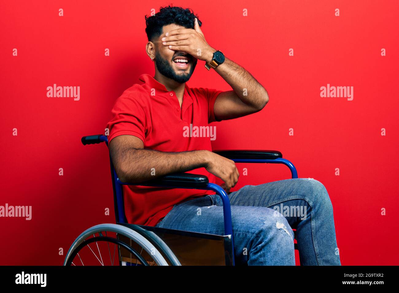 Uomo arabo con barba seduta su sedia a rotelle sorridente e ridendo con la  mano sulla faccia che copre gli occhi per sorpresa. Concetto cieco Foto  stock - Alamy