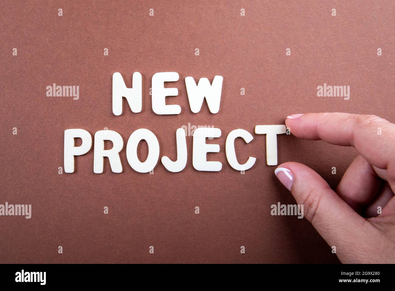 NUOVO PROGETTO. Lettere bianche di legno con testo e una mano di donna su sfondo marrone. Foto Stock