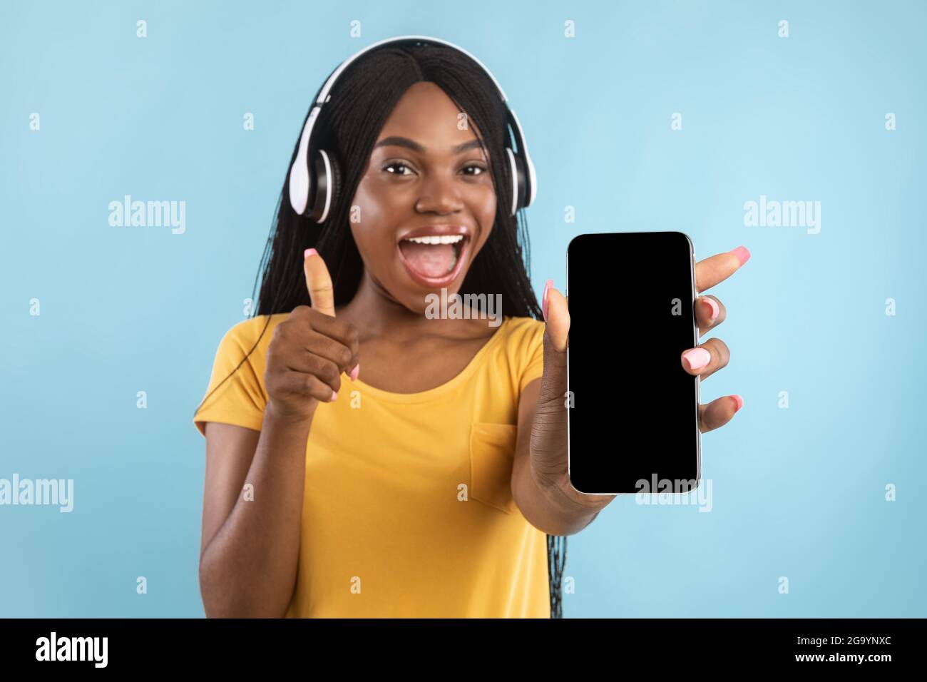 Donna nera che mostra lo schermo del telefono e Thumbs-Up che indossa le cuffie, studio Foto Stock