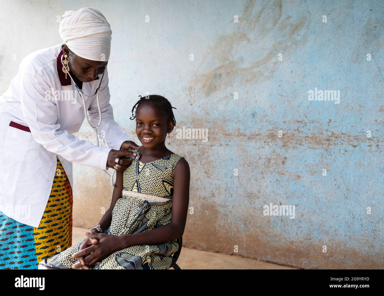 In questa immagine, un pediatra nero con un vestito da medico sta mettendo uno stetoscopio sul petto di una bambina sorridente con tipiche trecce africane du Foto Stock