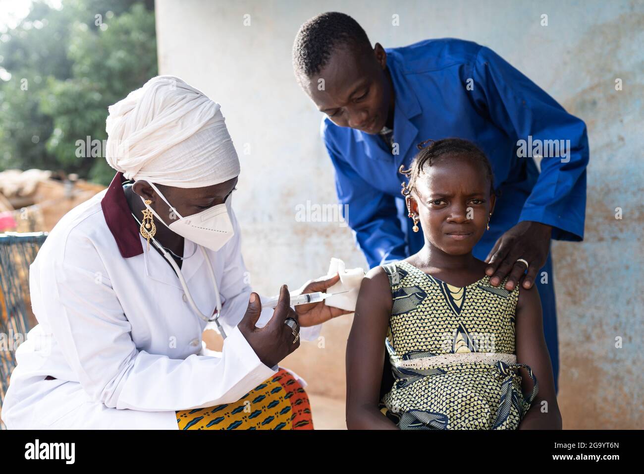 In questa immagine una ragazza africana nera spaventato che viene vaccinata da un'infermiera femminile con maschera facciale è confortata da un uomo in un cappotto blu che si piega ov Foto Stock