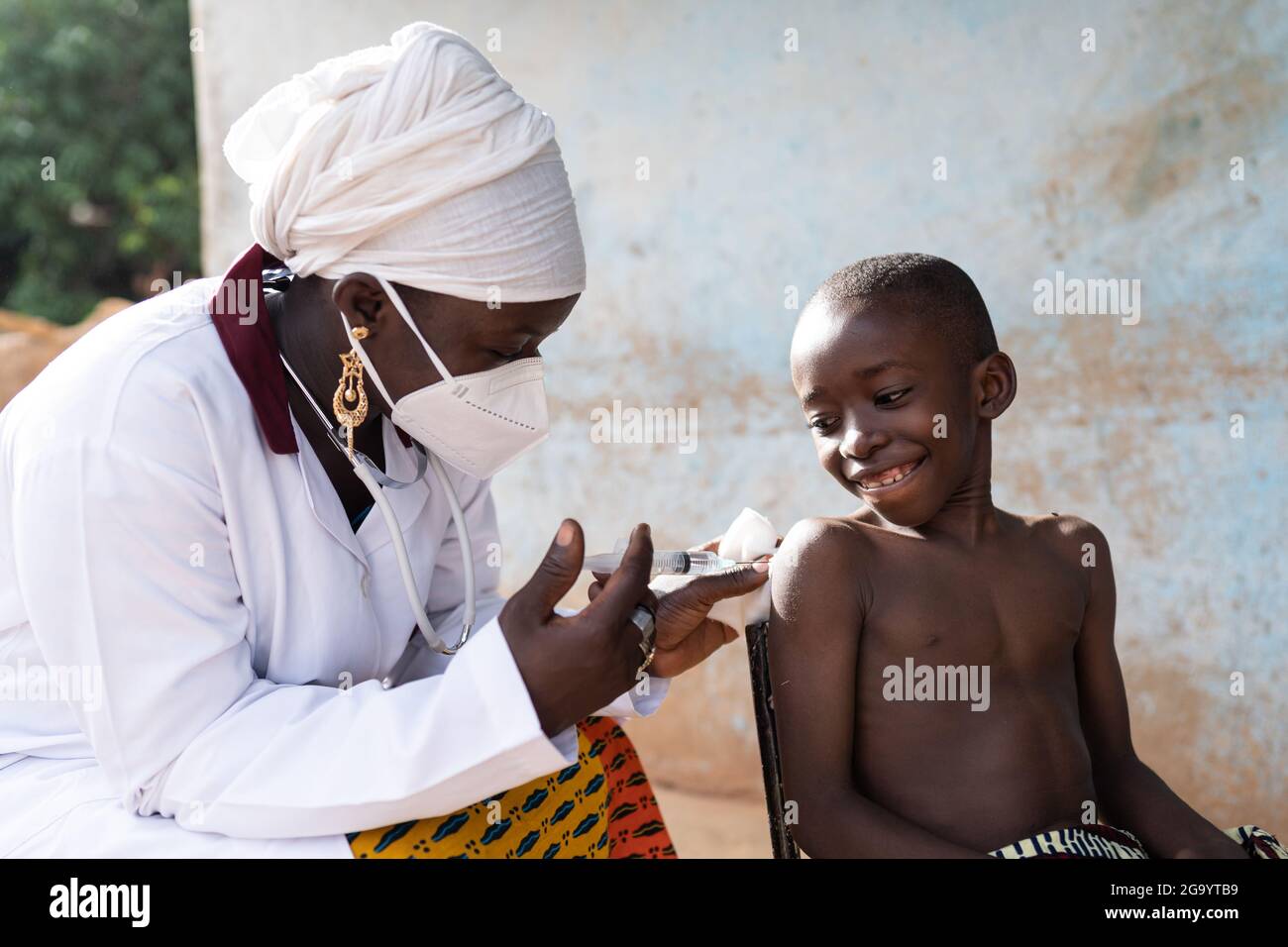 In questa immagine un'infermiera nera con maschera facciale sta iniettando una dose di vaccino ad un bravo studente sorridente in un ambiente ospedaliero africano Foto Stock