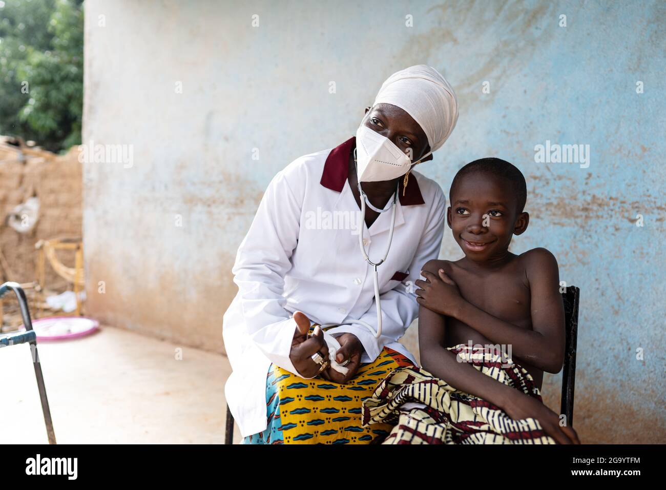 In questa immagine un piccolo ragazzo sorridente Afrian è seduto su una sedia che tiene il suo bastoncino di cotone sul braccio subito dopo essere stato vaccinato da un giovane BL Foto Stock