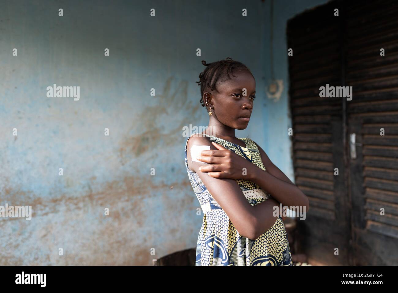 In questa immagine una piccola shoolgirl africana in un bel vestito estivo con un'espressione dolorosa sul suo viso è in piedi prima dell'ingresso di un decrepito rurale Foto Stock