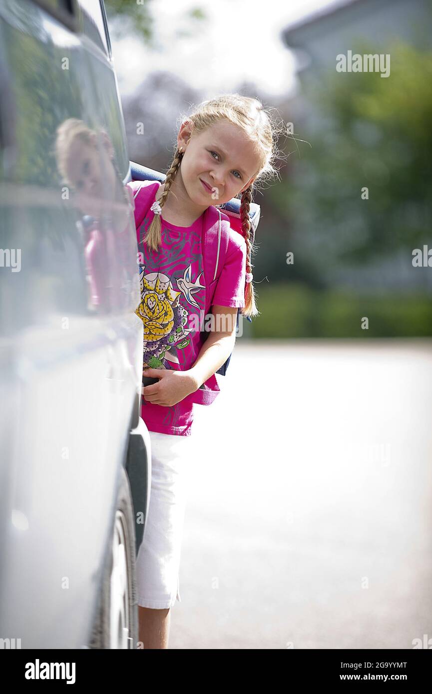 Felice studentessa elementare sbirciata dietro un'auto parcheggiata , Germania Foto Stock