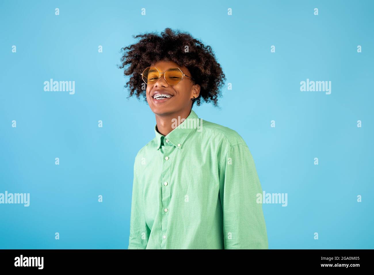 Ritratto di felice ragazzo teenager afroamericano in camicia di menta e occhiali da sole gialli che posano su sfondo blu studio Foto Stock