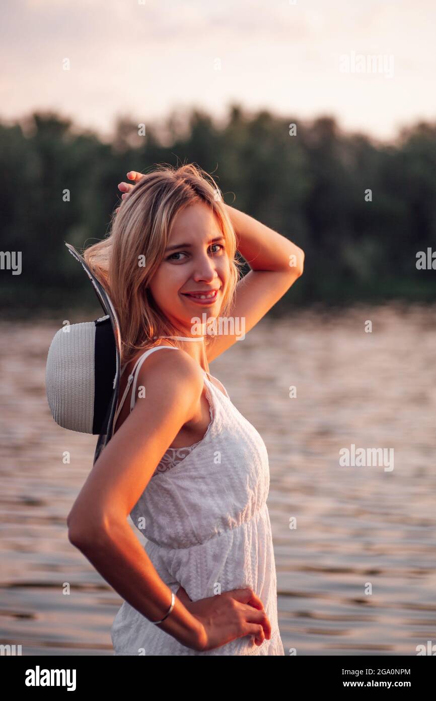 Ritratto di biondo sullo sfondo di un fiume. Una bella giovane donna felice posa al tramonto in un vestito bianco e un cappello di paglia con un nastro scuro Foto Stock