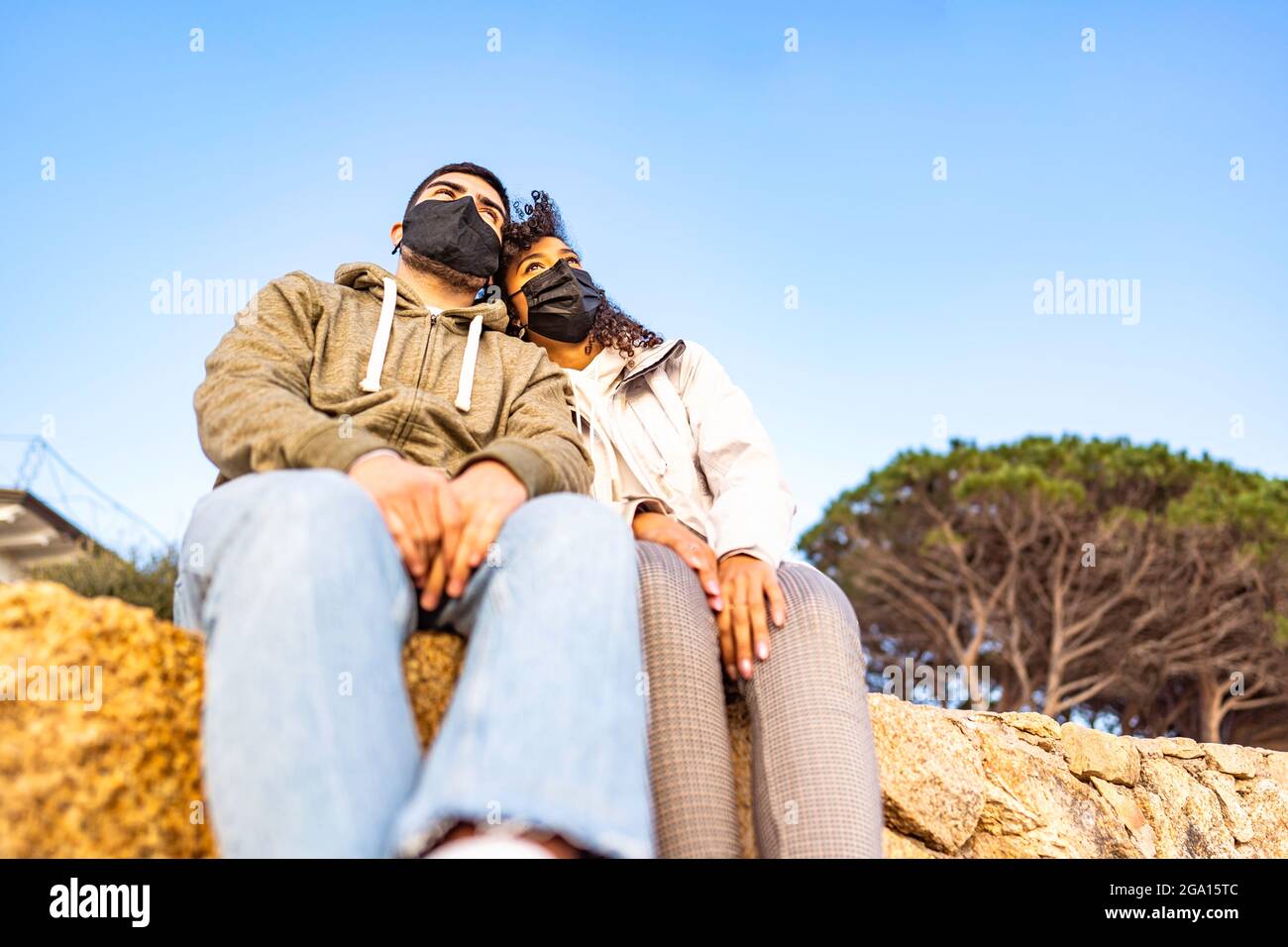 Giovane coppia multirazziale in amore seduto su una parete che indossa una maschera protettiva nera, testa a testa, guardando all'orizzonte al tramonto. Pandemia globale Foto Stock
