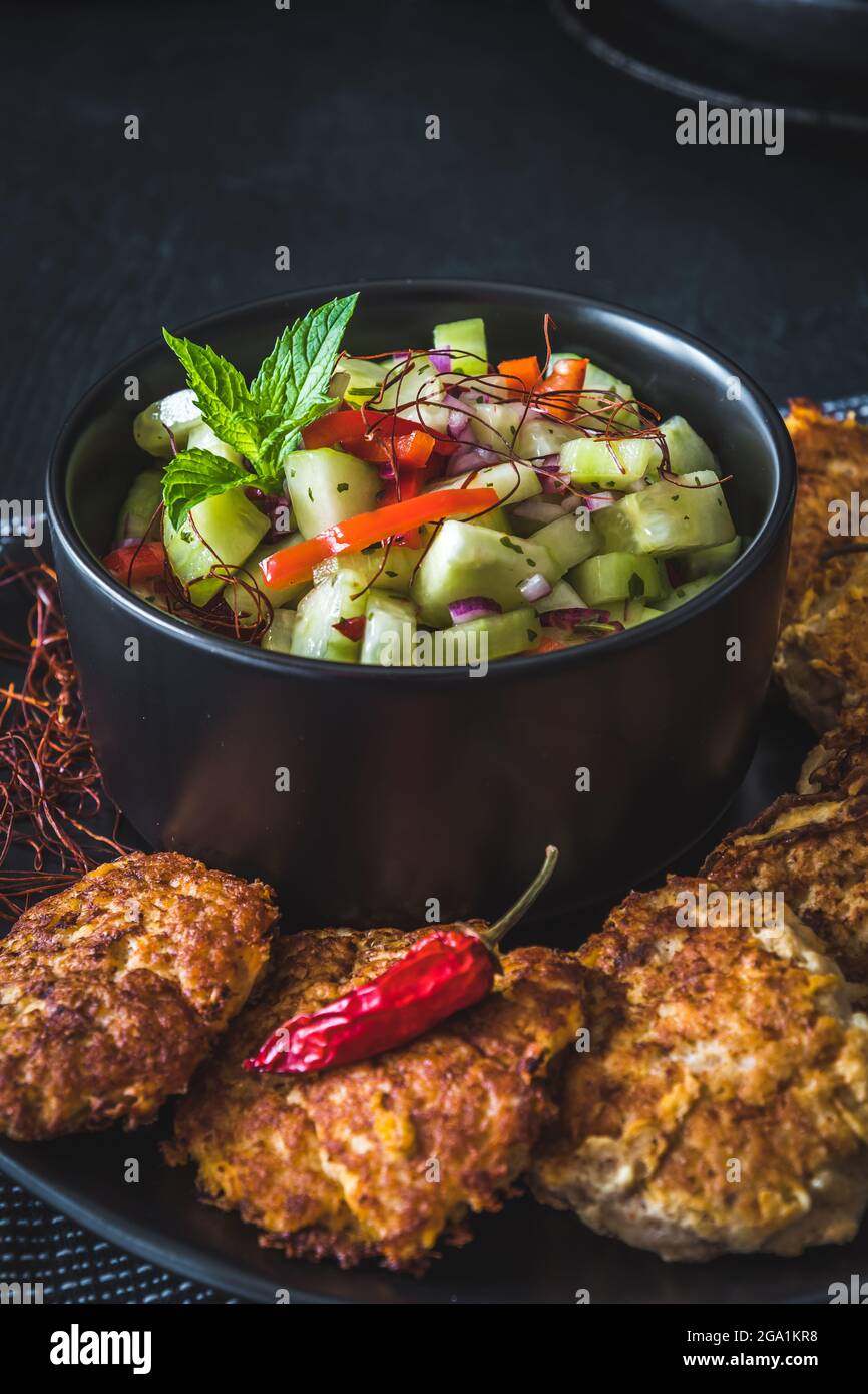 Insalata di cetrioli fresca in una ciotola nera con polpette di pesce o verdure o polpette di carne intorno, verticale Foto Stock